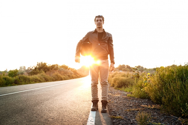 Junger schöner Motorradfahrer, der Helm hält und an Landstraße aufwirft.