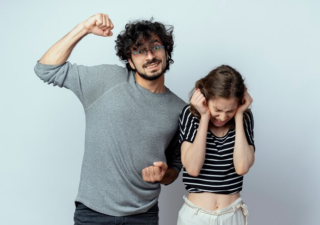 junger schöner Paarmann, der die Faust hebt, die neben seinem Mädchen steht und ihre Ohren mit den Händen mit genervtem Ausdruck über der weißen Wand schließt