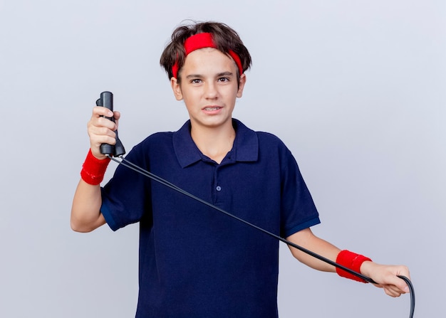 Junger schöner sportlicher Junge, der Stirnband und Armbänder mit Zahnspangen hält, die Springseil lokalisiert auf weißer Wand halten