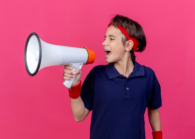 Junger schöner sportlicher Junge, der Stirnband und Armbänder mit Zahnspangen trägt, die Seite betrachten, die am Telefon lokalisiert auf purpurrotem Hintergrund spricht