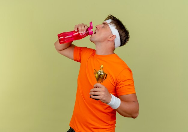 Junger schöner sportlicher Mann, der Stirnband und Armbänder hält, die Siegerbecher und Trinkwasser von der Wasserflasche lokalisiert auf olivgrünem Hintergrund mit Kopienraum halten