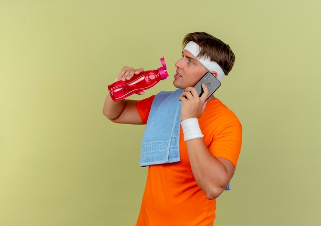 Kostenloses Foto junger schöner sportlicher mann, der stirnband und armbänder mit handtuch auf schulter spricht, die am telefon sprechen und wasserflasche lokalisiert auf olivgrünem hintergrund mit kopienraum halten