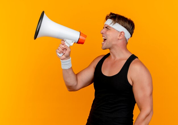 Junger schöner sportlicher Mann, der Stirnband und Armbänder schreit, die im Lautsprecher lokalisiert auf orange Hintergrund schreien
