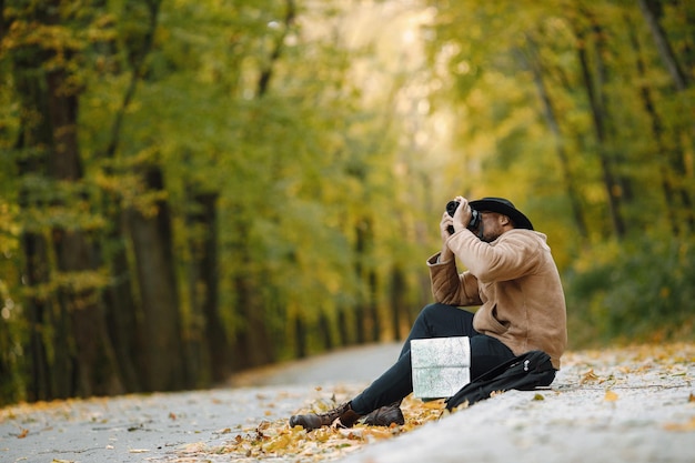 Junger schwarzer Mann, der mit einer Kamera auf der Straße im Wald sitzt. Männlicher Fotograf in einem Wald. Mann mit brauner Jacke, schwarzem Hut und Rucksack.