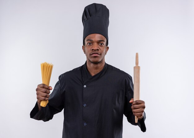 Junger selbstbewusster afroamerikanischer Koch in der Kochuniform hält Bündel von Spaghetti und Nudelholz lokalisiert auf weißem Hintergrund mit Kopienraum