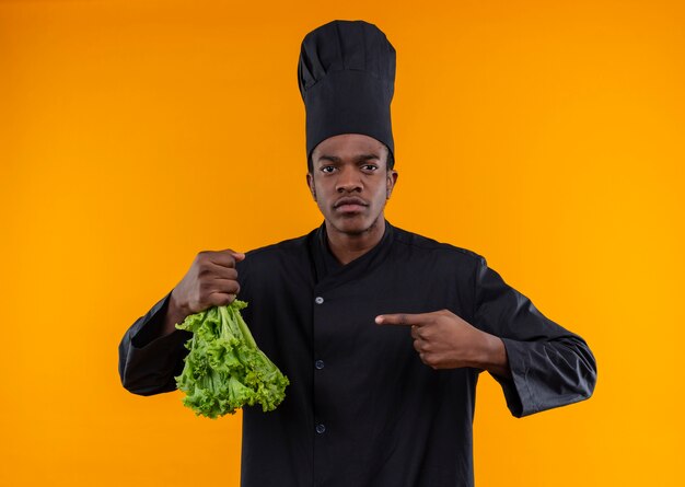 Junger selbstbewusster afroamerikanischer Koch in der Kochuniform hält Salat verkehrt herum isoliert auf orangefarbenem Hintergrund mit Kopienraum