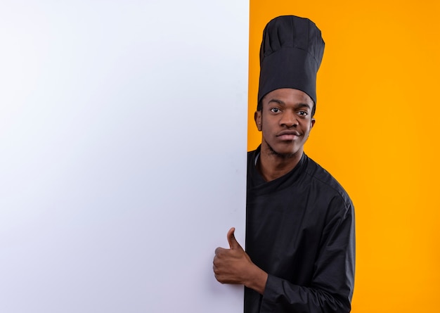 Junger selbstbewusster afroamerikanischer Koch in der Kochuniform steht hinter weißer Wand und Daumen hoch lokalisiert auf orange Hintergrund mit Kopienraum