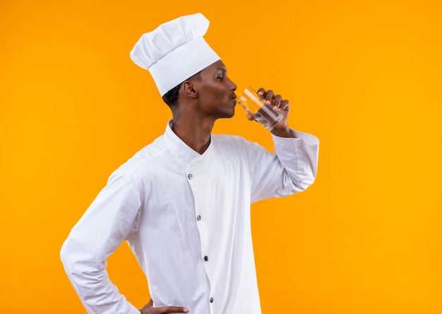 Junger selbstbewusster afroamerikanischer Koch in der Kochuniform steht seitlich und trinkt Glas Wasser lokalisiert auf orange Hintergrund mit Kopienraum