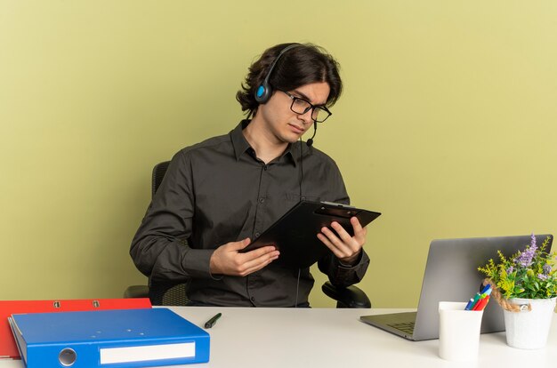 Junger selbstbewusster Büroangestellter Mann auf Kopfhörern in optischen Gläsern sitzt am Schreibtisch mit Bürowerkzeugen unter Verwendung von Laptop hält und betrachtet Zwischenablage lokalisiert auf grünem Hintergrund mit Kopienraum