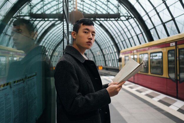 Junger seriöser asiatischer Geschäftsmann mit Zeitung, der selbstbewusst in die Kamera blickt, während er an der U-Bahn-Station auf den Zug wartet