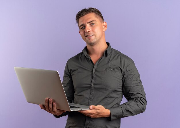 Junger sicherer blonder gutaussehender Mann hält Laptop lokalisiert auf violettem Raum mit Kopienraum