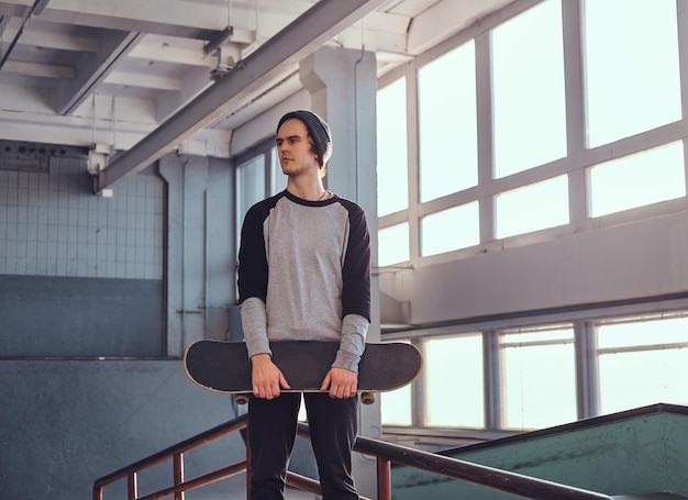 Junger Skateboarder, der drinnen neben einer Grindschiene im Skatepark steht, sein Board hält und wegschaut.