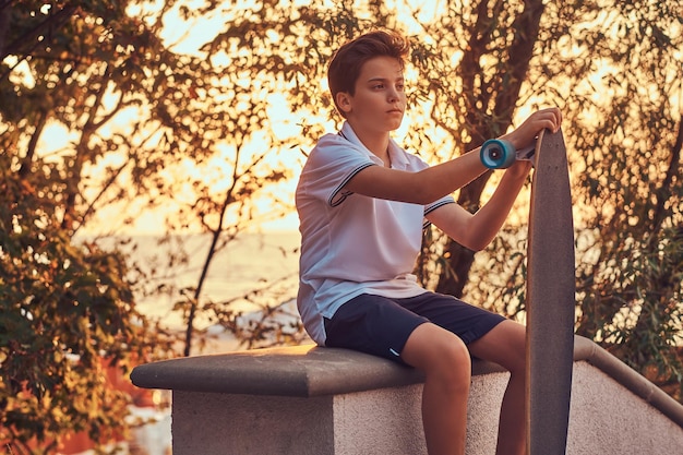 Junger Skater-Junge in T-Shirt und Shorts, der bei einem Sonnenuntergang auf der steinernen Leitplanke im Freien sitzt.