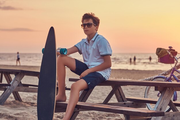 Junger Skater-Junge mit Sonnenbrille, gekleidet in T-Shirt und Shorts, sitzt auf einer Bank vor dem Hintergrund einer Meeresküste bei hellem Sonnenuntergang.