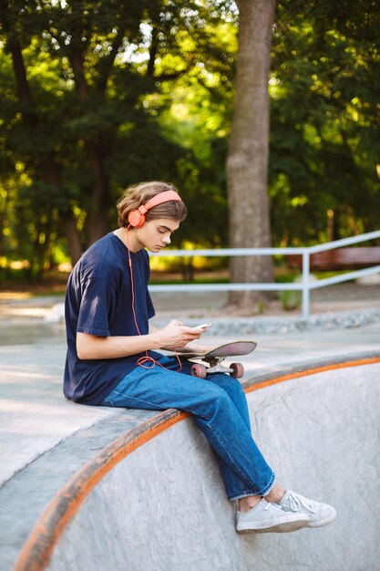 Junger Skater mit orangefarbenen Kopfhörern, während er nachdenklich Handy mit Skateboard in der Nähe eines modernen Skateparks benutzt