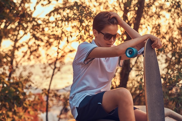 Junger skaterjunge mit sonnenbrille in t-shirt und shorts, der bei sonnenuntergang auf der steinleitplanke im freien sitzt.