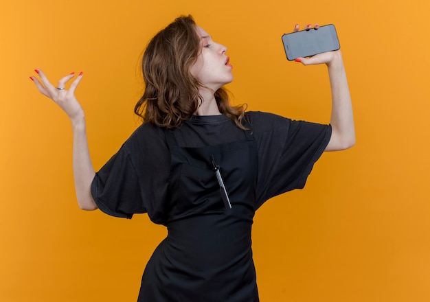 Junger slawischer weiblicher Friseur, der einheitliches Singen unter Verwendung des Mobiltelefons als Mikrofon trägt, das Hand in der Luft lokalisiert auf orange Hintergrund hält