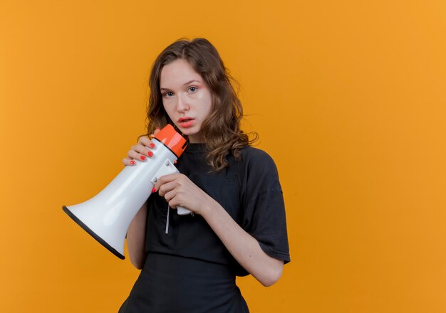 Junger slawischer weiblicher Friseur, der Uniform hält Lautsprecher hält, der Kamera lokalisiert auf orange Hintergrund mit Kopienraum betrachtet
