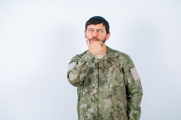 Junger soldat denkt, indem er eine haltende hand am kinn auf weißem hintergrund nachschlägt Premium Fotos