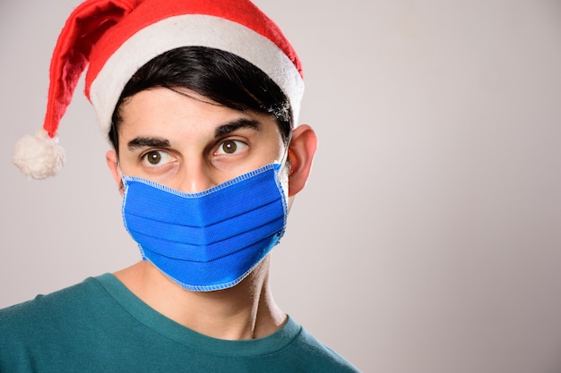 Junger spanischer Mann mit einer Gesichtsmaske und einer Weihnachtsmütze, die beiseite schaut