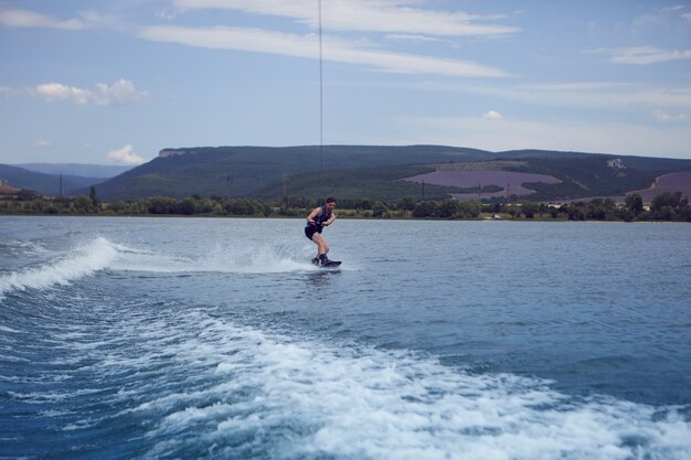 Junger Sportler, der über See surft. Surfer trägt nassen Badeanzug Training im Wake Park, Wakeboarden auf dem Fluss, von Motorboot gezogen, klammert sich an Kabel. Wakesurfen, Wasserski, Sport und Erholung