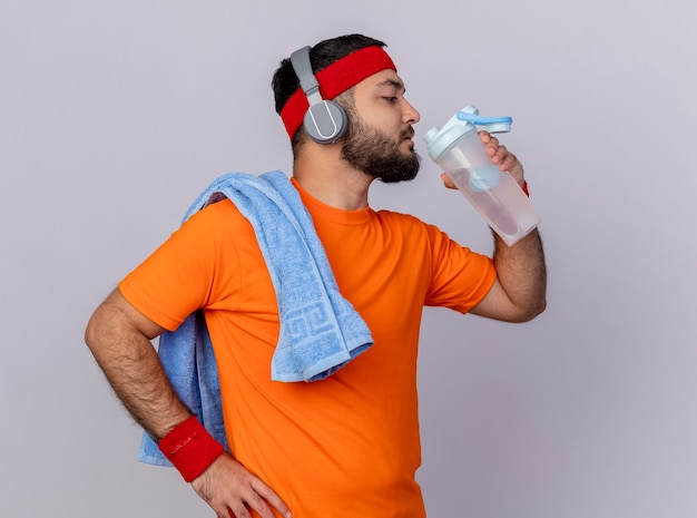 Junger sportlicher Mann, der in der Profilansicht das Tragen des Stirnbandes und des Armbandes mit Kopfhörern steht, trinkt Wasser von der Wasserflasche, die Hand auf Hüfte mit Handtuch auf Schulter lokalisiert auf weißem Hintergrund setzt