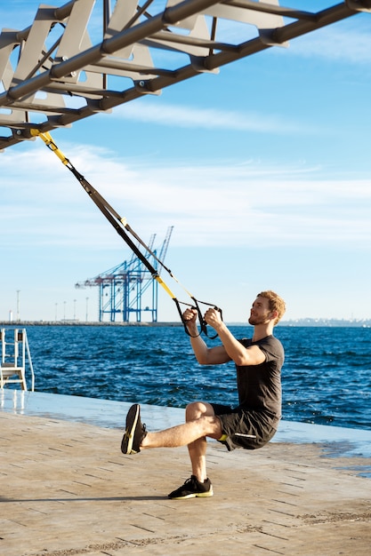 Junger sportlicher Mann, der mit trx nahe Meer am Morgen trainiert.