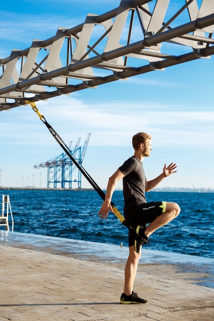 Junger sportlicher Mann, der mit trx nahe Meer am Morgen trainiert.