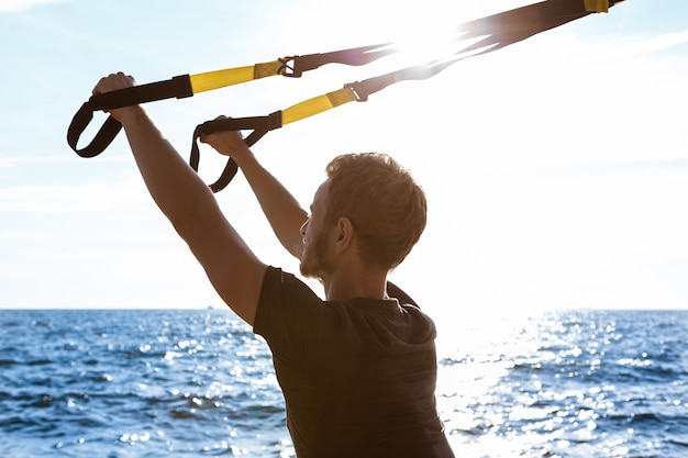 Kostenloses Foto junger sportlicher mann, der mit trx nahe meer am morgen trainiert.