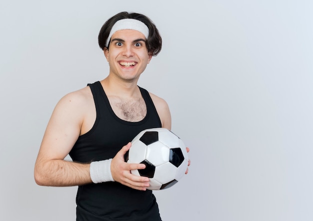 Junger sportlicher Mann, der Sportbekleidung und Stirnband hält Fußball hält, der vorne lächelnd mit glücklichem Gesicht steht über weißer Wand steht