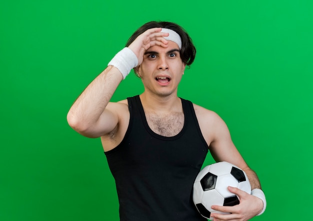 Junger sportlicher Mann, der Sportbekleidung und Stirnband hält Fußball hält mit Hand über Kopf verwechselt