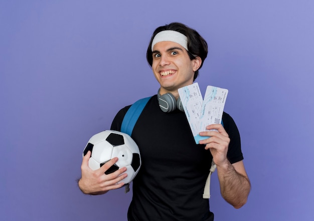 Junger sportlicher Mann, der Sportbekleidung und Stirnband mit Rucksack hält Fußball und Flugtickets lächelnd mit glücklichem Gesicht