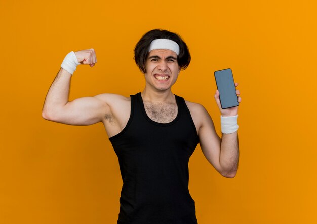 Junger sportlicher Mann, der Sportbekleidung und Stirnband trägt, das Smartphone zeigt, das Faust zeigt, die Bizeps zeigt, der angespannt aussieht