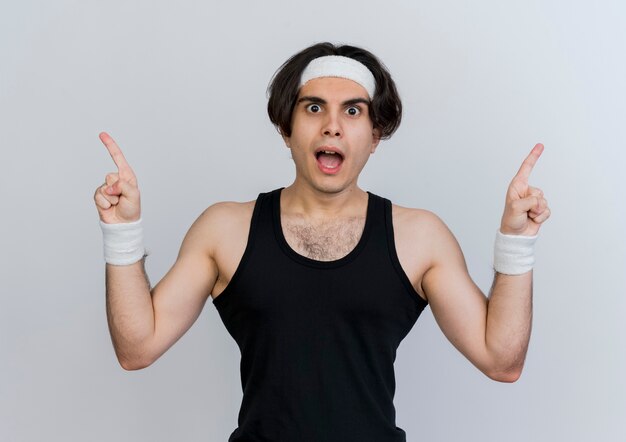 Junger sportlicher Mann, der Sportbekleidung und Stirnband trägt, die vorne überrascht mit Zeigefingern auf die Seiten schauen, die über weißer Wand stehen