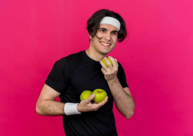 Junger sportlicher Mann, der Sportbekleidung und Stirnband trägt und grüne Äpfel lächelt, die fröhlich lächeln