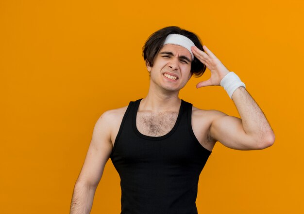 Junger sportlicher Mann, der Sportbekleidung und Stirnband trägt, verwirrt mit Hand auf seinem Kopf für Fehler