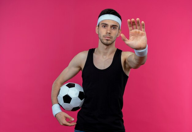 Junger sportlicher Mann im Stirnband, der Fußball hält Stoppschild mit offener Hand mit ernstem Gesicht, das über rosa Wand steht