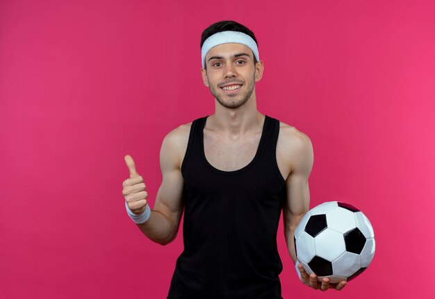 Junger sportlicher Mann im Stirnband, der Fußball lächelnd zeigt, zeigt Daumen hoch stehend über rosa Wand