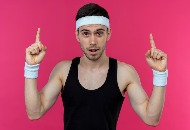 Junger sportlicher Mann im Stirnband, der Kamera mit sicherem Ausdruck betrachtet, der mit Zeigefingern oben steht über rosa Hintergrund zeigt