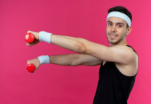 Junger sportlicher Mann im Stirnband, der mit Hanteln herausarbeitet, die glücklich und positiv über Rosa lächeln