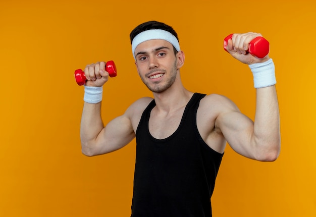 Junger sportlicher Mann im Stirnband, der mit Hanteln herausarbeitet, die über Orange lächeln