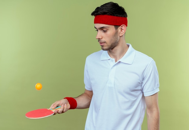 Junger sportlicher Mann im Stirnband, der Schläger für Tischtennis hält, der einen Ball wirft, der über grüner Wand steht