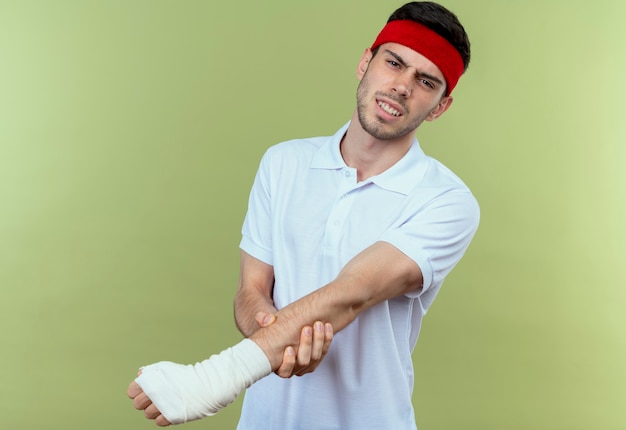 Junger sportlicher Mann im Stirnband, der seine bandagierte Hand berührt und Schmerz über Grün fühlt