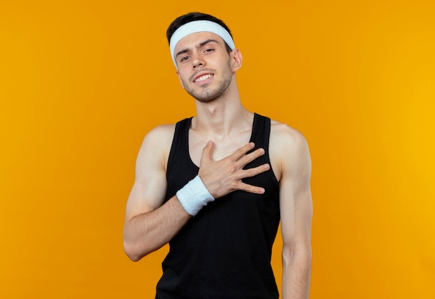 Junger sportlicher Mann im Stirnband, der seine Handfläche auf Brust hält, dankbar dankbar, über orange Wand stehend
