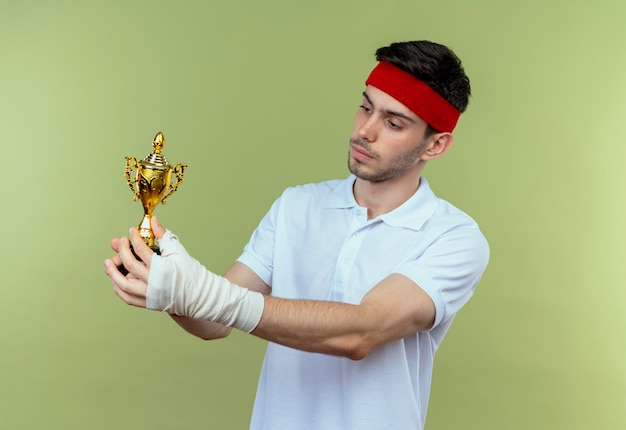 Kostenloses Foto junger sportlicher mann im stirnband, der seine trophäe hält, die sie mit sicherem ausdruck betrachtet, der über grüner wand steht