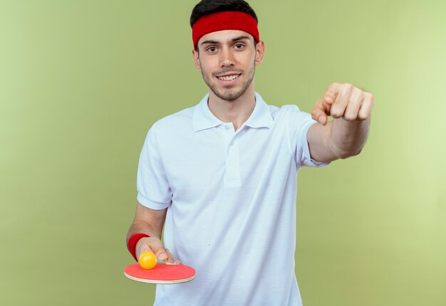 Junger sportlicher Mann im Stirnband hält Schläger und Ball für Tischtennis lächelnd mit Zeigefinger auf Sie stehend über grüne Wand