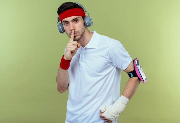 Junger sportlicher Mann im Stirnband mit Kopfhörern und Smartphone-Armband, die Stille Geste mit Finger auf Lippen über Grün machen