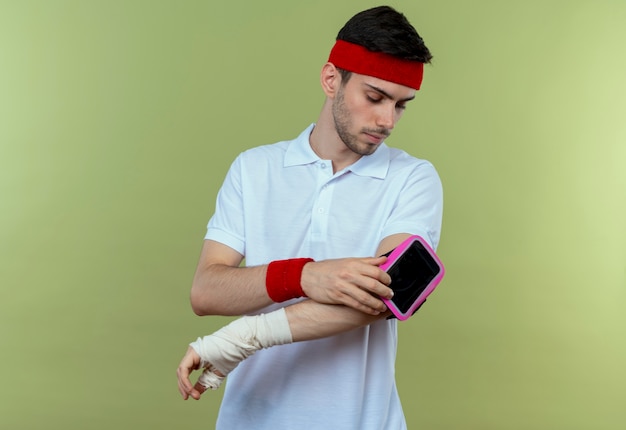 Junger sportlicher Mann im Stirnband mit Smartphone-Armband, das es mit ernstem Gesicht über Grün betrachtet