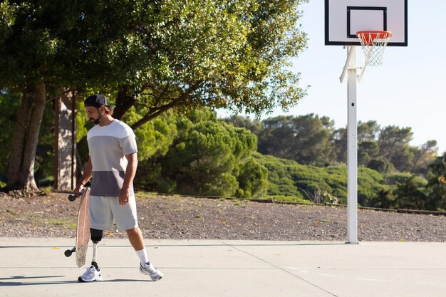 Junger sportlicher Mann mit Beinprothese, der den Basketballplatz verlässt, nachdem er dort Skateboard gefahren ist. Selbstbewusster bärtiger Typ mit Behinderung, der Skateboard hält, während er im Freien Sport treibt. Sportkonzept für Amputierte