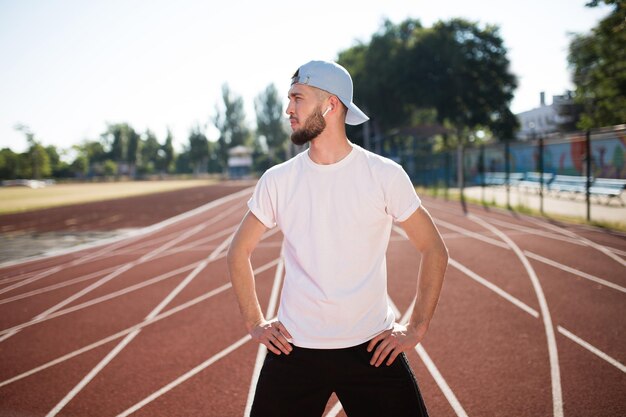 Junger sportlicher Mann mit drahtlosen Kopfhörern und weißer Kappe, der verträumt zur Seite schaut, während er Zeit auf dem Laufband des Stadions verbringt